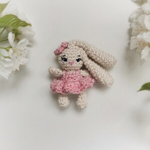 May include: A handmade crochet bunny doll with a pink dress and bow. The bunny has long ears, black eyes, and a pink nose. The doll is beige and pink, and is surrounded by white flowers.