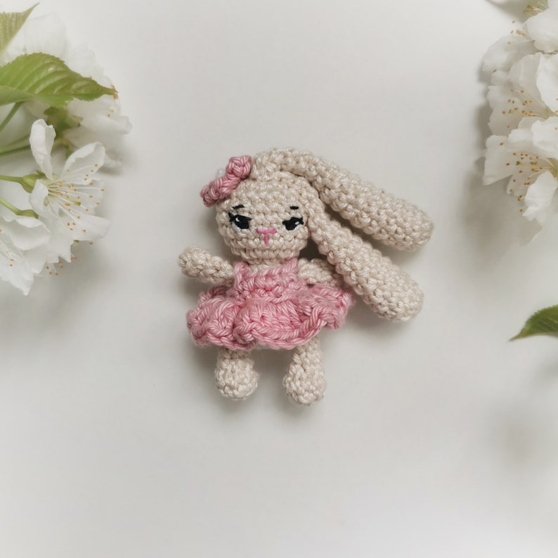 May include: A handmade crochet bunny doll with a pink dress and bow. The bunny has long ears, black eyes, and a pink nose. The doll is beige and pink, and is surrounded by white flowers.