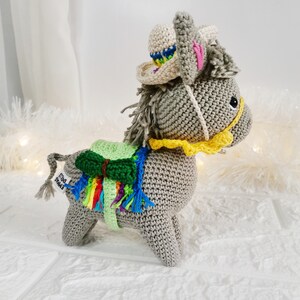 A crocheted grey donkey figurine wearing a sombrero and a colourful saddle blanket. The donkey has a yellow bridle and a grey mane and tail. The sombrero is beige with a colourful brim. The donkey is standing on a white surface.