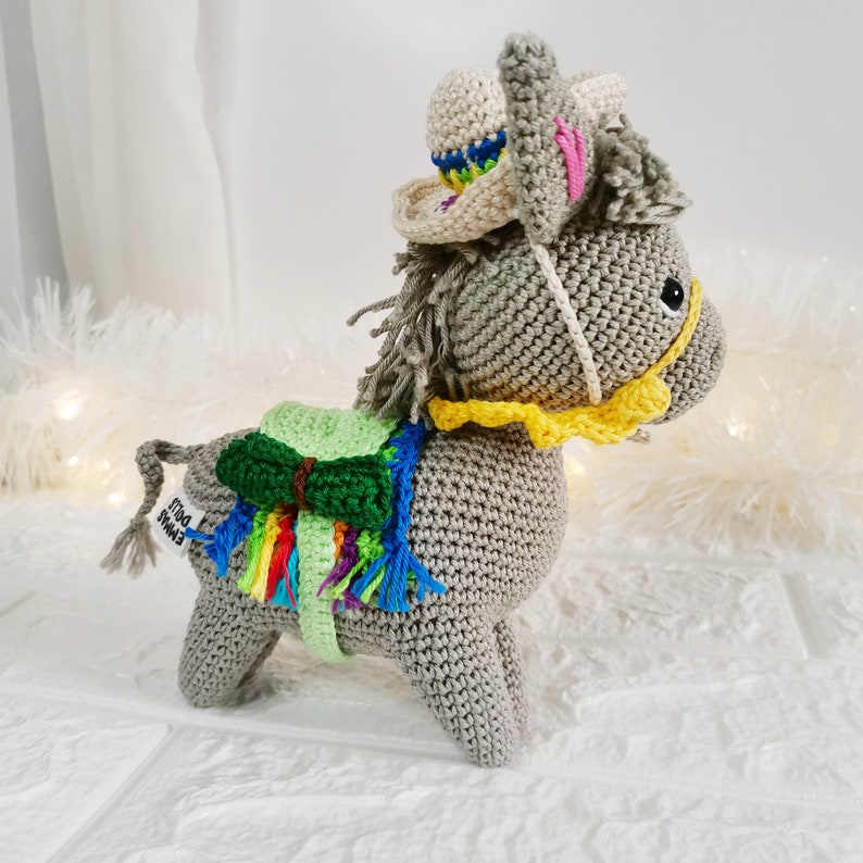 A crocheted grey donkey figurine wearing a sombrero and a colourful saddle blanket. The donkey has a yellow bridle and a grey mane and tail. The sombrero is beige with a colourful brim. The donkey is standing on a white surface.