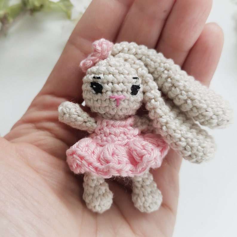 May include: A handmade crochet bunny doll with a pink dress and bow. The bunny has long, floppy ears, black eyes, and a pink nose. The doll is small and appears to be made of soft yarn. The overall aesthetic is cute and whimsical.