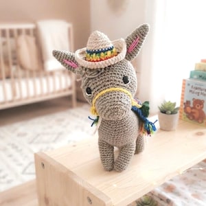 Festive donkey crochet pattern finished toy sitting on a wooden shelf with a mini sombrero and colorful fringed saddle.