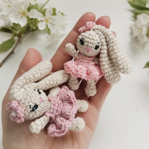 May include: Two crocheted bunny dolls. One bunny is lying down, and the other is standing. Both bunnies are beige with pink accents, including dresses and bows. The bunnies have black eyes and pink noses. The background is a hand holding the bunnies.