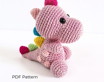 May include: A crocheted dinosaur toy in light pink with a dark pink crest, black plastic eyes, and a tail featuring small spikes in teal, yellow, light green, and dark pink. The body has light pink circular patches. Text reads: PDF Pattern.