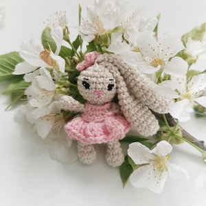May include: A handmade crochet bunny doll with a pink dress and bow, set against a backdrop of white blossoms and green leaves. The bunny has long, floppy ears and a sweet, embroidered face.