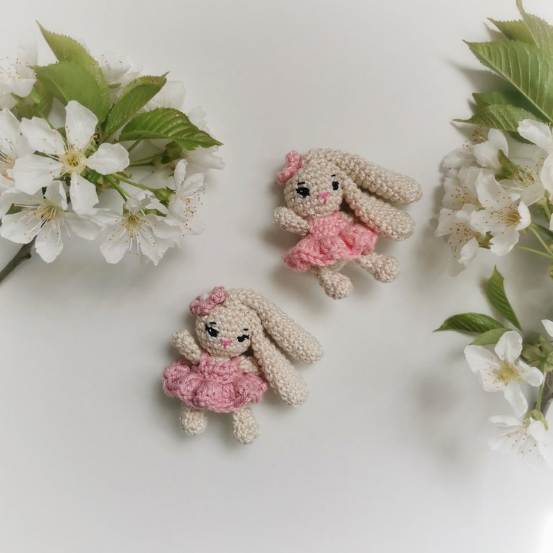 May include: Two handmade crochet bunny dolls. Each bunny has long ears, a pink dress, and a matching bow. The bunnies are beige and pink, and are surrounded by white flowers and green leaves.