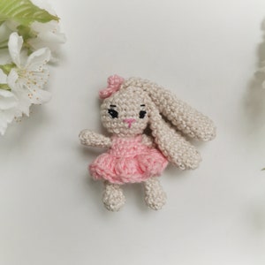 May include: A handmade crochet bunny doll with a pink dress and bow. The bunny has long ears, black eyes, and a pink nose. The doll is made of beige yarn and is set against a white background with white flowers.
