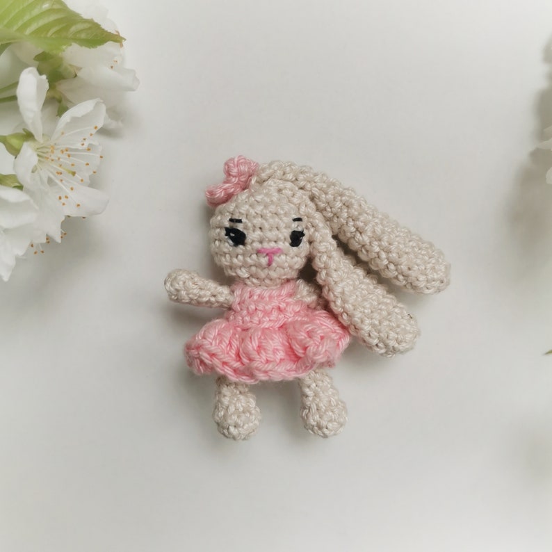 May include: A handmade crochet bunny doll with a pink dress and bow. The bunny has long ears, black eyes, and a pink nose. The doll is made of beige yarn and is set against a white background with white flowers.