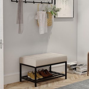 Può includere: Una panca imbottita beige con struttura in metallo nero e un ripiano inferiore per riporre le scarpe. La panca si trova in un corridoio con una porta bianca e un appendiabiti con una sciarpa e un cestino appesi.