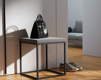 May include: A black leather handbag with gold-toned hardware sits on a square stool featuring a light gray upholstered seat and a black metal frame. The stool has four straight legs connected by a thin metal base, providing a stable structure.
