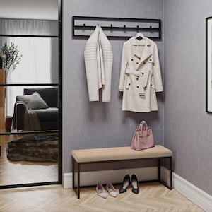 Könnte beinhalten: Eine schwarze Wandgarderobe aus Metall mit sechs Haken, an der eine weiße Jacke und ein beiger Trenchcoat hängen. Darunter steht eine beige gepolsterte Bank mit schwarzem Metallrahmen auf einem Holzboden, mit einer rosa Handtasche und zwei Paar Schuhen.