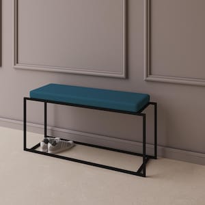 Op de afbeelding: Een moderne bank met een rechthoekig teal kussen en een zwart metalen frame. De bank heeft een onderste plank en een paar grijze sneakers staan op de plank. De bank staat tegen een grijze muur.