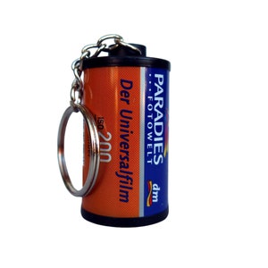 Op de afbeelding: Een sleutelhanger in de vorm van een filmrol met een zilveren sleutelring. De filmrol is oranje en blauw met de tekst "PARADIES FOTOWELT" en "Der Universalfilm". De film is ISO 200.
