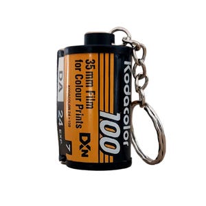 Puede incluir: Un llavero negro y naranja con forma de carrete de película Kodacolor de 35 mm. El carrete de película tiene el texto "Kodacolor 100" y "35 mm Film for Colour Prints DXN". El llavero incluye una cadena y un anillo plateados.