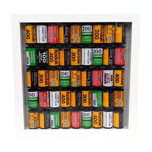 Könnte beinhalten: Ein weißer Rahmen, gefüllt mit einer Vielzahl von 35-mm-Filmkassetten. Die Kassetten sind in Reihen und Spalten angeordnet und zeigen verschiedene Marken, darunter Kodak, Fujicolor und Ilford. Die Kassetten sind alle in verschiedenen Farben, darunter Rot, Gelb, Grün und Blau.
