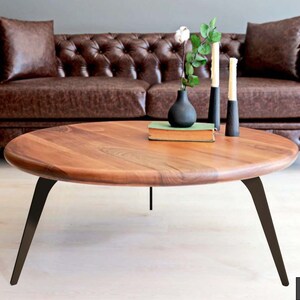 May include: A round wooden coffee table with a natural wood finish and black metal legs. The table is sitting in front of a brown leather sofa.