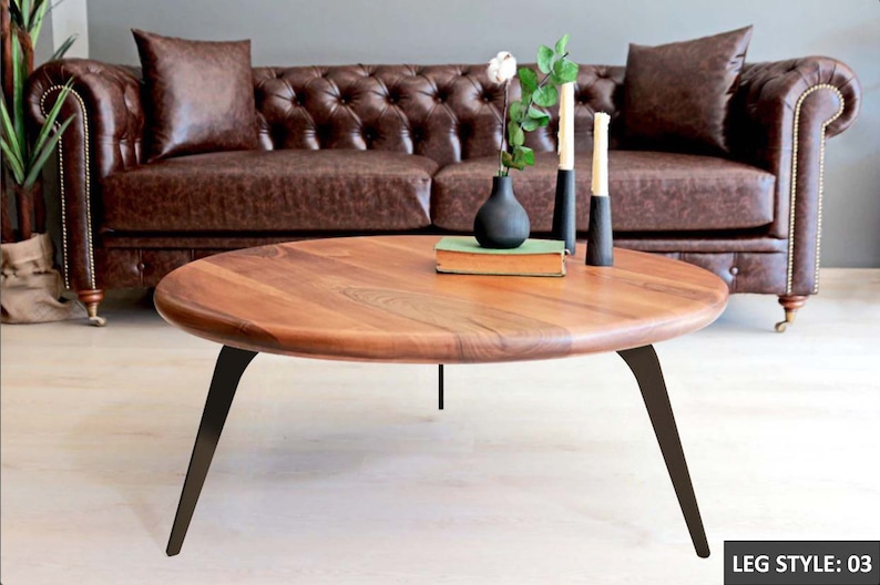 May include: A round wooden coffee table with a natural wood finish and black metal legs. The table is sitting in front of a brown leather sofa.