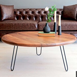 May include: A round wooden coffee table with a natural wood finish and black hairpin legs. The table is sitting in front of a brown leather sofa.