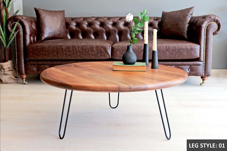 May include: A round wooden coffee table with a natural wood finish and black hairpin legs. The table is sitting in front of a brown leather sofa.