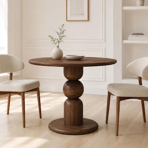 May include: A round dark wood dining table with a central pedestal base composed of three stacked spherical and cylindrical shapes, accompanied by two light beige upholstered chairs with wooden legs, set in a bright room.