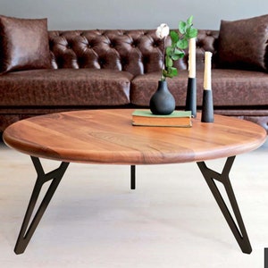 May include: A round wooden coffee table with a natural wood finish and black metal legs. The table has a unique leg style with a Y-shaped design. The table is sitting in front of a brown leather sofa.