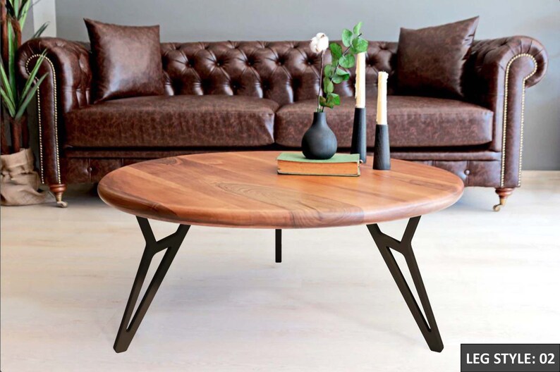 May include: A round wooden coffee table with a natural wood finish and black metal legs. The table has a unique leg style with a Y-shaped design. The table is sitting in front of a brown leather sofa.