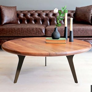 May include: A round wooden coffee table with a natural wood finish and black metal legs. The table has a unique, slightly curved design and is sitting in front of a brown leather sofa.