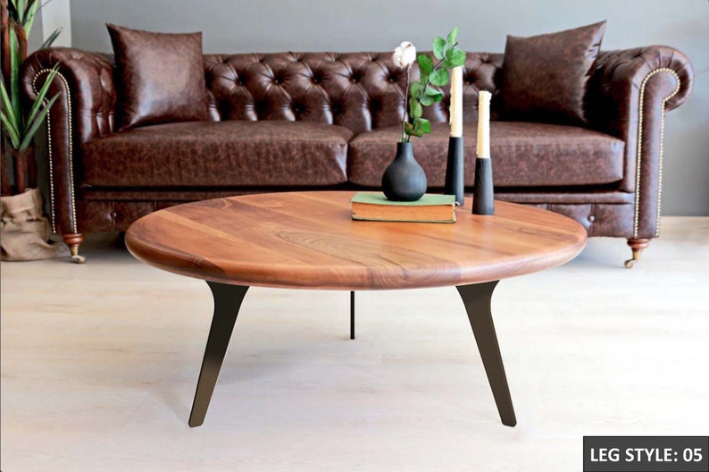 May include: A round wooden coffee table with a natural wood finish and black metal legs. The table has a unique, slightly curved design and is sitting in front of a brown leather sofa.