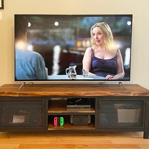 May include: A dark wood TV stand with metal mesh doors and shelves. The stand has two doors and two shelves. The top shelf is empty, the middle shelf has a black video game console and the bottom shelf has two green and pink video game controllers. A flat screen TV is sitting on top of the stand.
