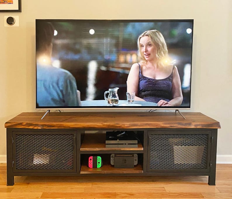 May include: A dark wood TV stand with metal mesh doors and shelves. The stand has two doors and two shelves. The top shelf is empty, the middle shelf has a black video game console and the bottom shelf has two green and pink video game controllers. A flat screen TV is sitting on top of the stand.