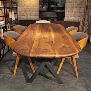 Puede incluir: Una mesa de comedor de madera oscura con una forma única, sostenida por una base de metal negro. La mesa está rodeada por seis sillas con marcos de madera y asientos tapizados en gris. La habitación tiene una paleta de colores neutros.