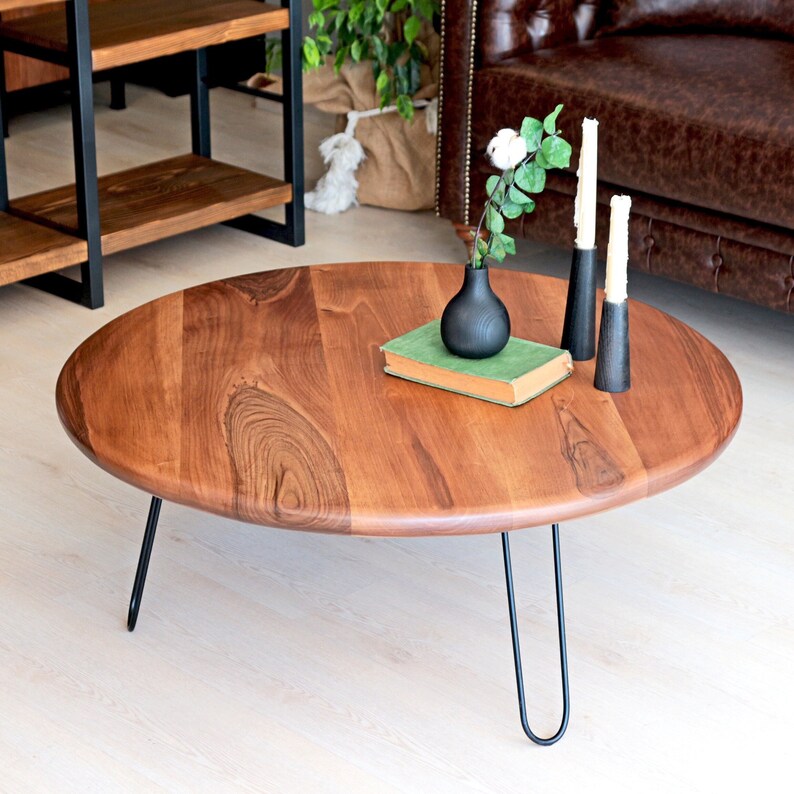 May include: A round wooden coffee table with black hairpin legs. The table has a natural wood finish and is decorated with a book, a vase with a green plant, and three candlesticks.