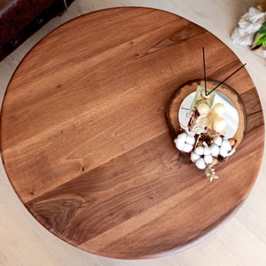May include: A round wooden table top with a natural wood grain finish. The table top is decorated with a small decorative tray with white cotton balls, gold ornaments, and a glass jar with gold ornaments.