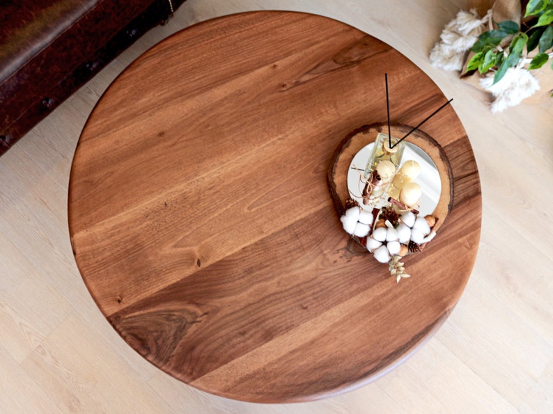 May include: A round wooden table top with a natural wood grain finish. The table top is decorated with a small decorative tray with white cotton balls, gold ornaments, and a glass jar with gold ornaments.