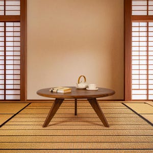 May include: A round wooden coffee table with a natural wood finish sits on a straw mat. The table has a simple design with three legs and a wide, flat top. A white teapot and a cup and saucer are on the table.