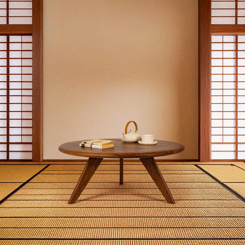May include: A round wooden coffee table with a natural wood finish sits on a straw mat. The table has a simple design with three legs and a wide, flat top. A white teapot and a cup and saucer are on the table.