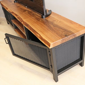 May include: A wooden and metal TV stand with a live edge top and black metal mesh doors. The doors are open, revealing a shelf inside.