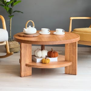 以下が含まれることがあります： 丸い木製のコーヒーテーブル。上部には白いティーポット、2つのティーカップ、木製のサービングボードがあります。下の棚には、白、オレンジ、黄色の装飾的なニットカボチャが飾られています。
