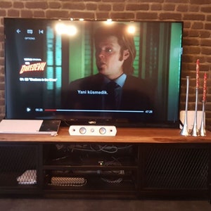May include: A home entertainment system with a large flat screen TV, a sound bar, and two white tower speakers. The TV is playing a movie with the title "Daredevil" on the screen. The speakers are on a black metal and wood entertainment stand.