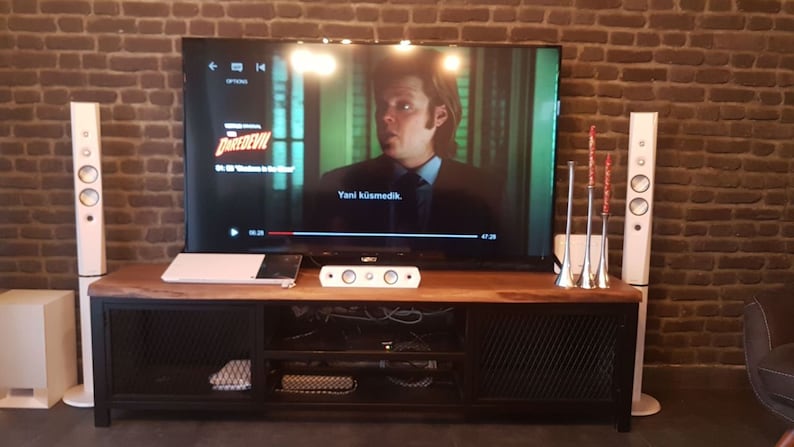 May include: A home entertainment system with a large flat screen TV, a sound bar, and two white tower speakers. The TV is playing a movie with the title "Daredevil" on the screen. The speakers are on a black metal and wood entertainment stand.