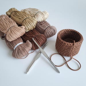 Puede incluir: Un surtido de suministros de ganchillo, que incluye varios ovillos de hilo beige, marrón y canela, dos ganchillos plateados y una pequeña cesta redonda de ganchillo marrón. El hilo está cuidadosamente empaquetado con bandas elásticas blancas.