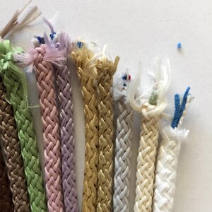 May include: Assortment of braided cords in various colors, including brown, green, pink, lavender, gold, gray, cream, and white. The cords are all approximately the same thickness and have a slightly textured surface.
