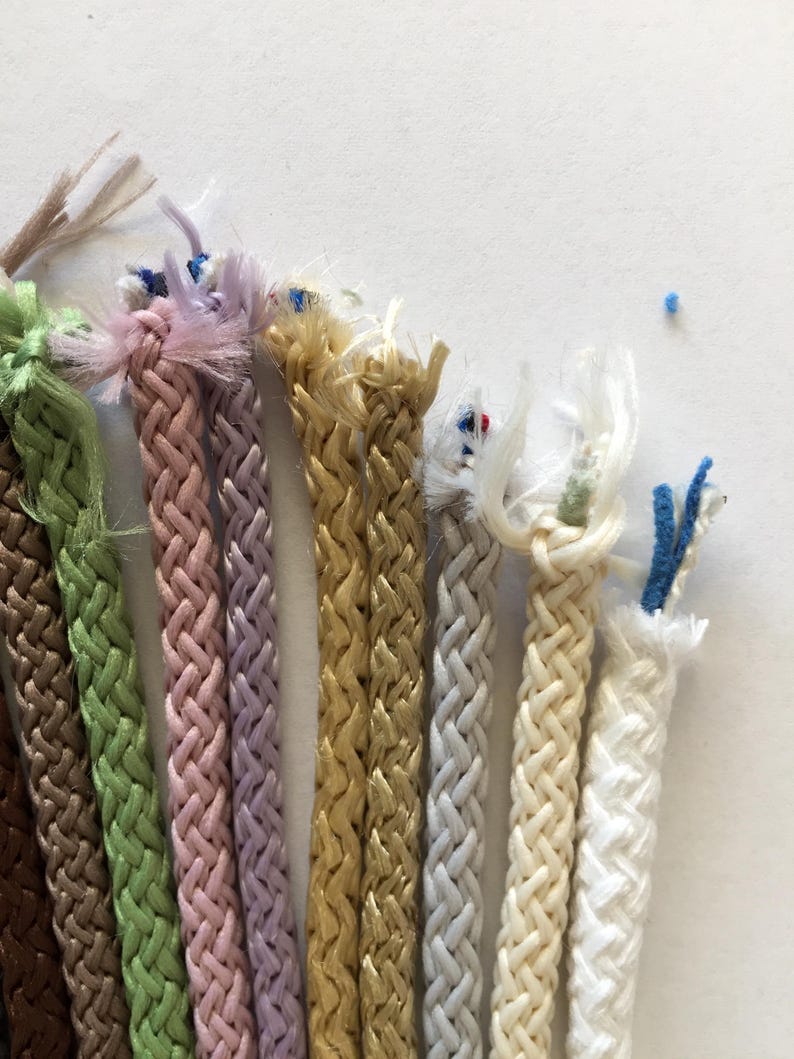 May include: Assortment of braided cords in various colors, including brown, green, pink, lavender, gold, gray, cream, and white. The cords are all approximately the same thickness and have a slightly textured surface.