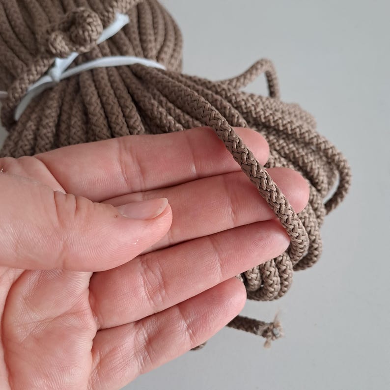 May include: A close-up of a spool of brown cotton cord. The cord is thick and has a braided texture. The cord is being held in a hand.