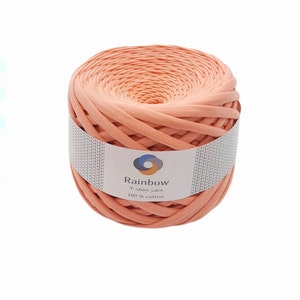 May include: A large, tightly wound ball of peach-colored Rainbow T-shirt yarn, made of 100% cotton. The yarn is wrapped with a white label featuring the brand name and logo. Ideal for various crafting projects.