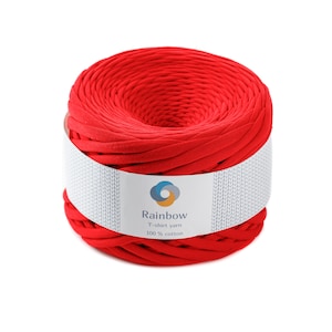 May include: A vibrant red T-shirt yarn, tightly wound into a cylindrical shape. A white label wraps around the yarn, displaying the brand name "Rainbow" and the text "T-shirt yarn 100% cotton". The yarn is a solid, bright red color.
