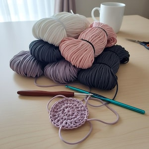 May include: A collection of yarn balls in various colors, including white, pink, gray, and purple, arranged on a light-colored surface. A crochet project in progress with a lilac-colored circle is visible, along with two crochet hooks. A white mug is in the background.