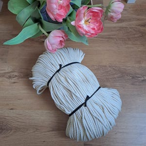 May include: A bundle of cream-coloured rope, secured with black ties, lies on a wooden surface. Pink tulips and green leaves are arranged in the background. The rope appears to be made of a woven material.