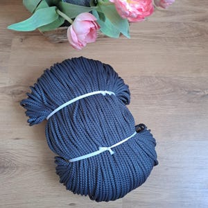 Peut inclure: Une grande bobine de corde en coton gris foncé. La corde est enroulée serrée et fixée avec un ruban en plastique blanc.