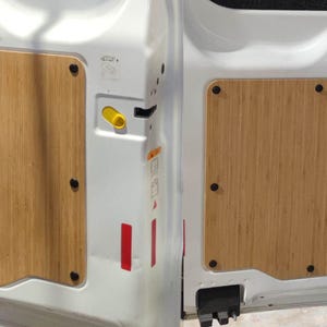 May include: Interior van door panels with bamboo-style wood inserts. The panels are rectangular and secured with black fasteners. The van doors are white, and the panels are a light brown color. The door has a yellow handle and red reflective stickers.